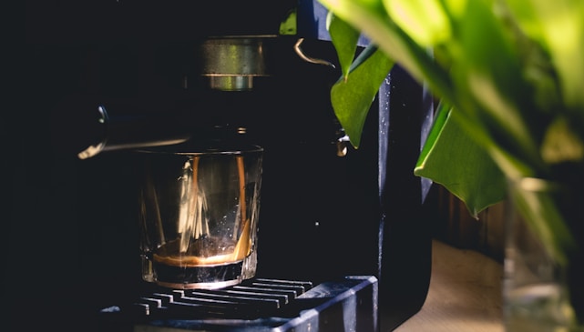 A coffee machine with a glass cup collecting freshly brewed espresso. The machine is partially obscured by green leaves, creating a contrast between the mechanical elements and the natural foliage.