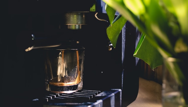 A coffee machine with a glass cup collecting freshly brewed espresso. The machine is partially obscured by green leaves, creating a contrast between the mechanical elements and the natural foliage.