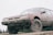 A rugged car covered in mud is parked on a grassy terrain, featuring large, textured tires suitable for off-road driving. The vehicle appears to have been used in a muddy environment, with dirt caked onto the sides and undercarriage.
