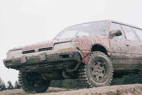 A rugged car covered in mud is parked on a grassy terrain, featuring large, textured tires suitable for off-road driving. The vehicle appears to have been used in a muddy environment, with dirt caked onto the sides and undercarriage.