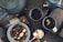 A rustic tea setting features two dark ceramic cups filled with tea on a wooden surface. A matching teapot sits nearby, partially visible. A stone bowl contains loose leaf tea with various dried fruits and petals. A spoon of dried flowers and a woven fabric enhance the cozy, natural ambiance.