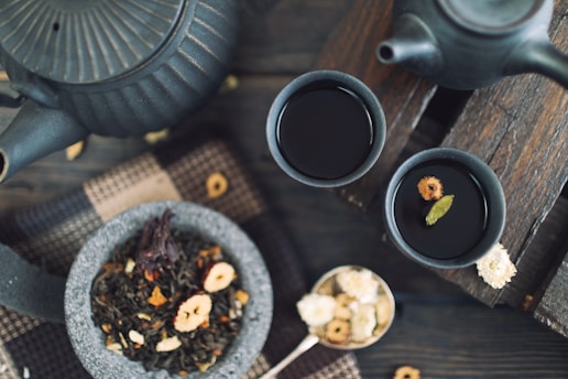 A rustic tea setting features two dark ceramic cups filled with tea on a wooden surface. A matching teapot sits nearby, partially visible. A stone bowl contains loose leaf tea with various dried fruits and petals. A spoon of dried flowers and a woven fabric enhance the cozy, natural ambiance.