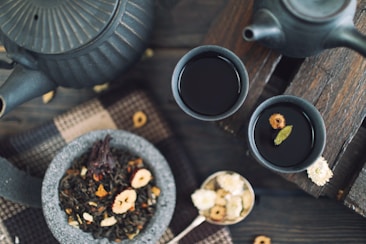 A rustic tea setting features two dark ceramic cups filled with tea on a wooden surface. A matching teapot sits nearby, partially visible. A stone bowl contains loose leaf tea with various dried fruits and petals. A spoon of dried flowers and a woven fabric enhance the cozy, natural ambiance.