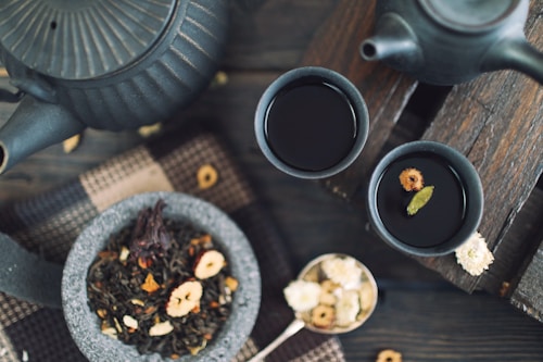 A rustic tea setting features two dark ceramic cups filled with tea on a wooden surface. A matching teapot sits nearby, partially visible. A stone bowl contains loose leaf tea with various dried fruits and petals. A spoon of dried flowers and a woven fabric enhance the cozy, natural ambiance.