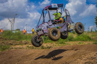 A racing vehicle with an open frame design is airborne during a dirt track event. The driver, equipped with a helmet and protective gear, is navigating the rough terrain. In the background, people in bright, colorful clothes are visible, watching the race under a blue sky with scattered clouds.