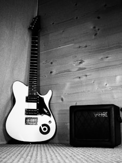 A white electric guitar with black accents and a logo rests against a wooden wall beside a black Vox amplifier on a textured carpet.