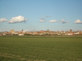 A village with a mix of old and modern buildings sits under a sky scattered with fluffy clouds. The foreground is dominated by a lush green field, and various earthy tones of the village's rooftops contrast against the clear blue sky.