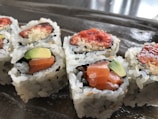 Close-up view of sushi rolls with visible ingredients like salmon, avocado, and a crispy topping. The rolls are arranged on a reflective surface, highlighting the texture of the rice and fillings.