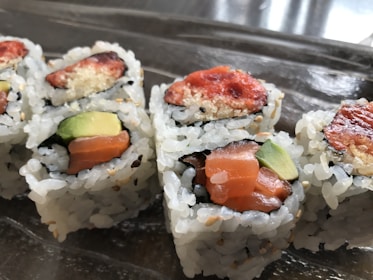 Close-up view of sushi rolls with visible ingredients like salmon, avocado, and a crispy topping. The rolls are arranged on a reflective surface, highlighting the texture of the rice and fillings.