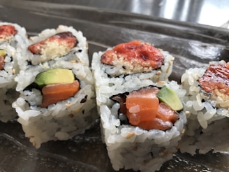 Close-up view of sushi rolls with visible ingredients like salmon, avocado, and a crispy topping. The rolls are arranged on a reflective surface, highlighting the texture of the rice and fillings.