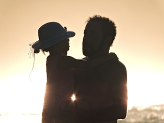 Silhouette of an adult and child embracing against a bright, backlit sunset. The child is wearing a hat, and both figures appear in profile.