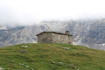 A small stone cabin stands on a grassy hillside with a backdrop of majestic, rugged mountains partially shrouded in mist. The landscape is serene and remote, creating a sense of tranquility and isolation.