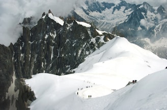 Snow-covered mountains with dramatic, jagged peaks and a line of climbers traversing the ridge. Clouds partially obscure the view, adding a mysterious atmosphere to the rugged landscape.