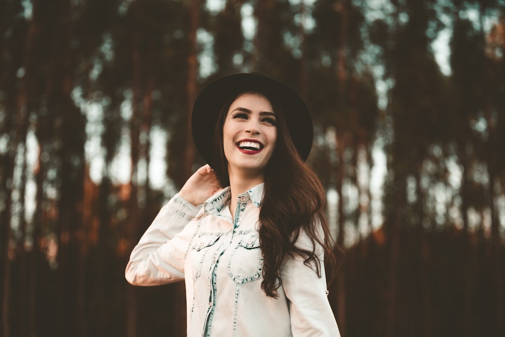 A woman in dark lipstick and a black hat smiles broadly in front of tall trees