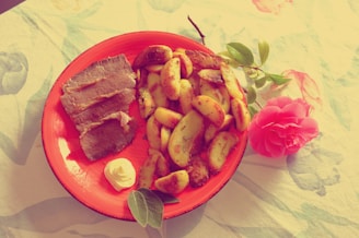 A vibrant plate of roasted potato wedges and slices of meat on a red ceramic plate, accompanied by a small dollop of creamy sauce. A pink flower lies beside the plate on a table covered with a floral-patterned tablecloth, highlighting a rustic and homely setting.