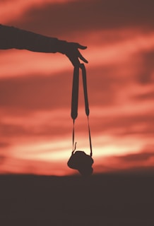 A silhouette of a hand holding a camera by its strap against a vibrant sunset sky. The sky features warm hues of red, orange, and pink blending together, creating a dramatic and peaceful atmosphere. The outline of the camera adds a sense of storytelling to the scene.