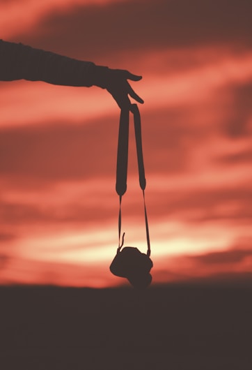 A silhouette of a hand holding a camera by its strap against a vibrant sunset sky. The sky features warm hues of red, orange, and pink blending together, creating a dramatic and peaceful atmosphere. The outline of the camera adds a sense of storytelling to the scene.