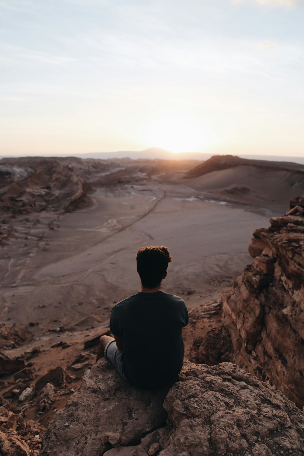 man sitting on cliff facing open field during daytime