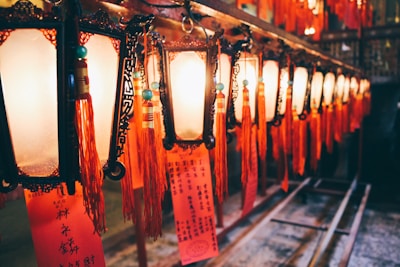 A series of ornate, traditional lanterns hang in a row, each illuminated and adorned with red tassels and written notes or prayers. The warm glow of the lights casts a cozy ambiance, while the intricate designs on the lanterns add to the cultural aesthetic.