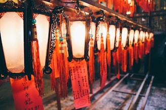 A series of ornate, traditional lanterns hang in a row, each illuminated and adorned with red tassels and written notes or prayers. The warm glow of the lights casts a cozy ambiance, while the intricate designs on the lanterns add to the cultural aesthetic.