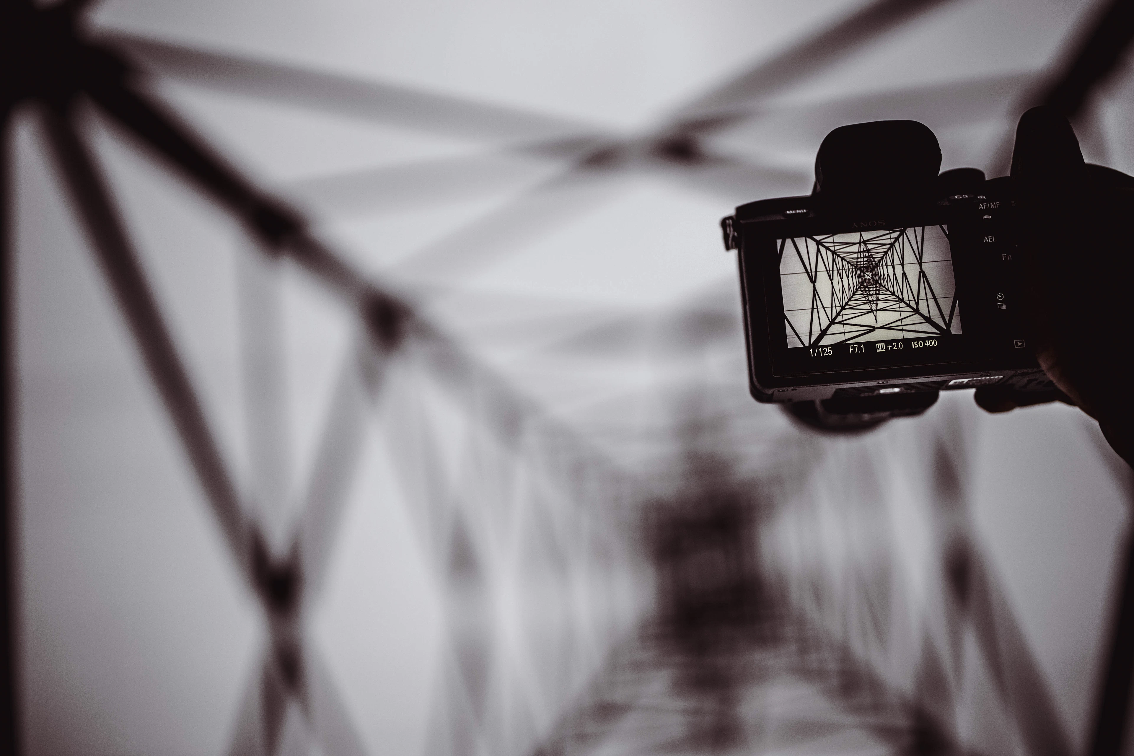 The rear screen of a camera held up against a steel lattice tower, framing the structure from below in black and white