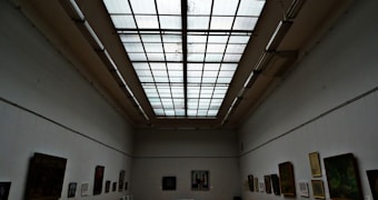 A spacious art gallery with a high ceiling features large rectangular skylights. The walls are lined with various paintings and artworks, each framed and hung neatly. The atmosphere is quiet and contemplative, with a focus on the displayed art.