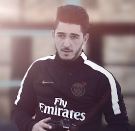 A man with styled hair and facial hair is wearing a black sports jacket with white stripes and yellow accents. The jacket displays logos, including one that reads 'Paris' and another with the word 'Emirates.' He holds a camera in his hands and looks slightly off to the side with a neutral expression. The background is blurred, suggesting an outdoor setting with warm lighting.