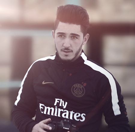 A man with styled hair and facial hair is wearing a black sports jacket with white stripes and yellow accents. The jacket displays logos, including one that reads 'Paris' and another with the word 'Emirates.' He holds a camera in his hands and looks slightly off to the side with a neutral expression. The background is blurred, suggesting an outdoor setting with warm lighting.