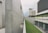 A modern architectural structure featuring minimalistic concrete walls and linear geometric forms. The design incorporates clean lines and open spaces, with a blend of greenery including a narrow grass strip along the edge of the walkway. One structure has a grid-like facade while another incorporates extensive windows. The surrounding scenery includes a mountain partially obscured by the buildings.