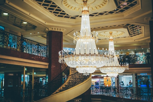 A luxurious interior featuring an ornate chandelier hanging from the ceiling, surrounded by intricate wrought-iron railings and a grand staircase. The setting is elegant with shops visible in the background, adorned with detailed architectural elements and soft lighting.