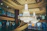 A luxurious interior featuring an ornate chandelier hanging from the ceiling, surrounded by intricate wrought-iron railings and a grand staircase. The setting is elegant with shops visible in the background, adorned with detailed architectural elements and soft lighting.