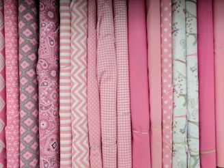 A collection of rolls of fabric, primarily in shades of pink and white. The fabrics feature various patterns, including anchors, paisley, chevrons, gingham, polka dots, and floral prints. All the rolls are neatly arranged in a row, showcasing their patterns and colors.