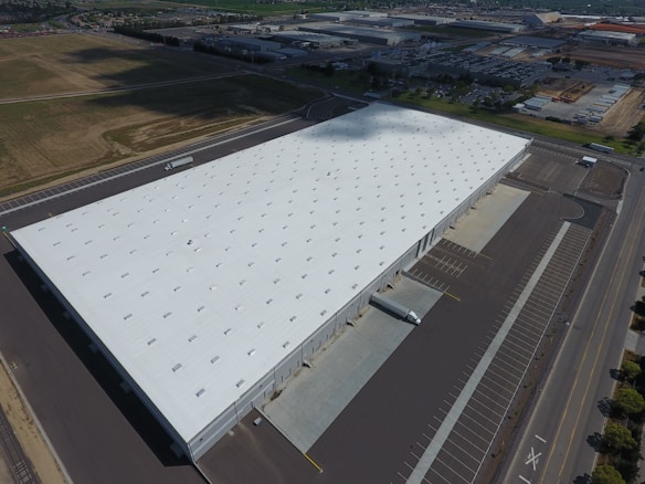 A large warehouse with a white roof dominates the landscape, surrounded by parking lots and vast open areas. The building is rectangular and appears to be part of an industrial complex. A few trucks, including a small white one, are parked nearby, indicating logistical activity.