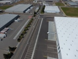 An aerial view of an industrial area with large warehouses, parking lots, and a road lined with trees. There are several parked trucks with trailers and some cargo containers near the buildings. One truck is parked next to a loading area of a large warehouse.