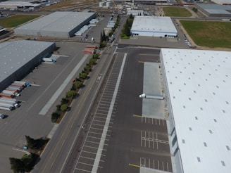 An aerial view of an industrial area with large warehouses, parking lots, and a road lined with trees. There are several parked trucks with trailers and some cargo containers near the buildings. One truck is parked next to a loading area of a large warehouse.