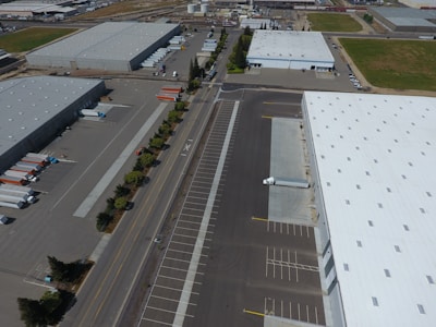 An aerial view of an industrial area with large warehouses, parking lots, and a road lined with trees. There are several parked trucks with trailers and some cargo containers near the buildings. One truck is parked next to a loading area of a large warehouse.