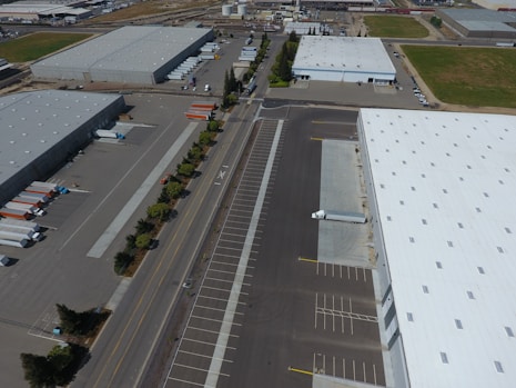 An aerial view of an industrial area with large warehouses, parking lots, and a road lined with trees. There are several parked trucks with trailers and some cargo containers near the buildings. One truck is parked next to a loading area of a large warehouse.
