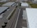 An aerial view of an industrial area with large warehouses, parking lots, and a road lined with trees. There are several parked trucks with trailers and some cargo containers near the buildings. One truck is parked next to a loading area of a large warehouse.