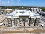 Aerial view of a modern residential building under construction with a flat rooftop, surrounded by other buildings and trees. Scaffolding is visible on the side, and construction materials are scattered around. The sky is clear with few clouds.