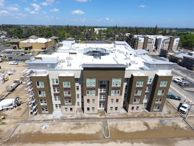 Aerial view of a modern residential building under construction with a flat rooftop, surrounded by other buildings and trees. Scaffolding is visible on the side, and construction materials are scattered around. The sky is clear with few clouds.