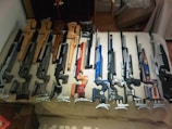 A collection of precision rifles are neatly arranged on a checkered surface. Each rifle features detailed craftsmanship with wooden and metallic elements, and they vary in color, predominantly showcasing shades of brown, black, blue, and red. The arrangement suggests they are either on display or being prepared for an event or gathering.