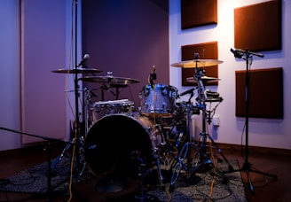 A drum set is placed in a soundproofed room with acoustic panels mounted on the walls. The drum kit includes cymbals, toms, and a snare drum, surrounded by several microphones on stands for recording. The lighting casts a warm glow, creating a cozy and professional atmosphere suitable for musical performances.