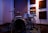 A drum set is placed in a soundproofed room with acoustic panels mounted on the walls. The drum kit includes cymbals, toms, and a snare drum, surrounded by several microphones on stands for recording. The lighting casts a warm glow, creating a cozy and professional atmosphere suitable for musical performances.