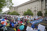 A large crowd of people is gathered in a street, many holding signs and banners advocating for science. The event is set against the backdrop of a grand, historical building with classical architecture and numerous windows. Participants are dressed in rain gear, and some are carrying umbrellas, indicating rainy weather. The atmosphere appears to be lively and spirited, with diverse and colorful attire.