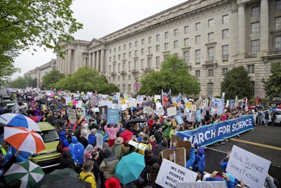 A large crowd of people is gathered in a street, many holding signs and banners advocating for science. The event is set against the backdrop of a grand, historical building with classical architecture and numerous windows. Participants are dressed in rain gear, and some are carrying umbrellas, indicating rainy weather. The atmosphere appears to be lively and spirited, with diverse and colorful attire.
