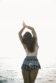 A person stands by the water with their arms raised above their head in a yoga pose. They are facing away, and the sunlight creates a serene, backlit effect. The person is wearing a light-colored top and patterned shorts, with long hair flowing down their back.