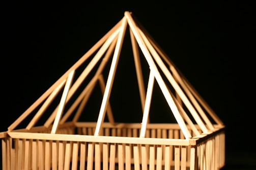A model of a house made from wooden sticks, featuring a triangle-shaped roof structure against a dark background.