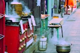 A street food stall with steaming pots and bamboo steamers, surrounded by a vibrant urban setting. The stall features a red counter with brass handles and hanging signs. A variety of cooking equipment and food items are visible, while a bicycle can be seen in the background.