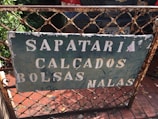 A weathered metal sign is affixed to a rusted fence, with the words 'Sapataria Calçados Bolsas Malas' in faded white paint. The background is a dark green color, with visible chipping and peeling. Behind the fence is a parked car with a visible license plate and green foliage on the left side.
