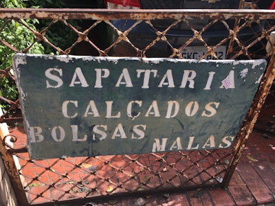 A weathered metal sign is affixed to a rusted fence, with the words 'Sapataria Calçados Bolsas Malas' in faded white paint. The background is a dark green color, with visible chipping and peeling. Behind the fence is a parked car with a visible license plate and green foliage on the left side.